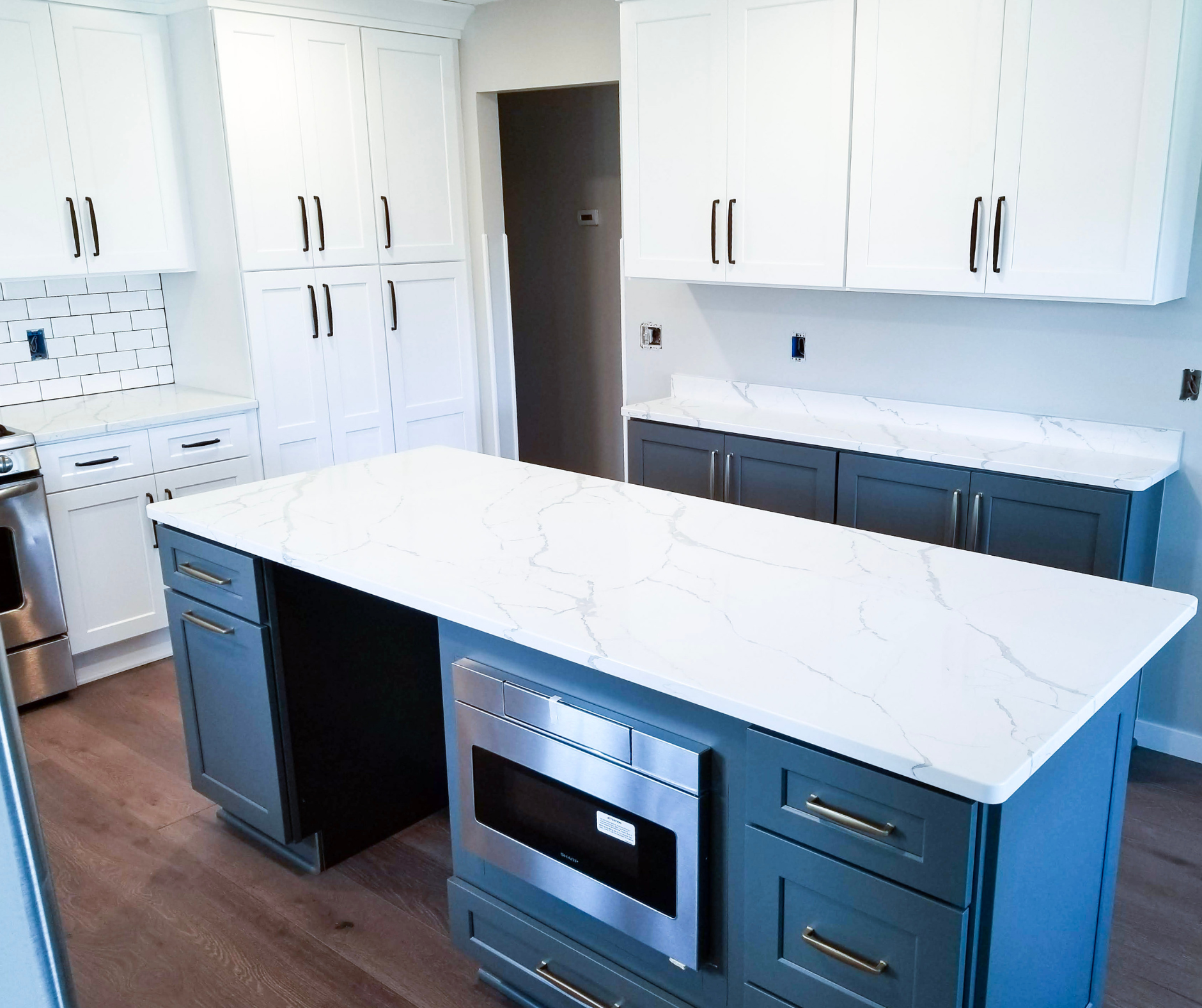 Beautiful, remodeled kitchen with blue lower cabinets, white upper cabinets, and white countertops. There is a large kitchen island with an oven in it and all hardware is modern and dark colored.